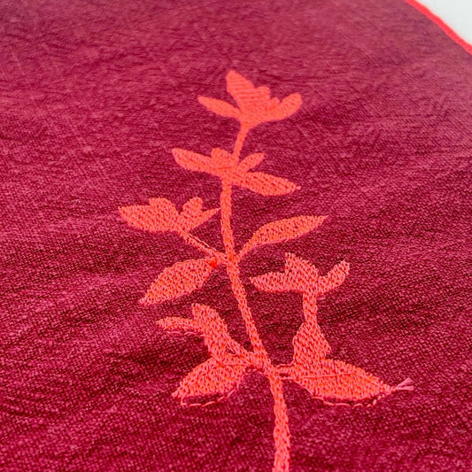 Serviette de table en lin lavé de couleur framboise, ornée de finitions en rose néon. Elle présente un motif brodé de thym, une plante aromatique. Vue du produit.