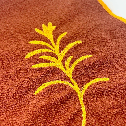Serviette de table en lin lavé de couleur piment, ornée de finitions en orange néon. Elle présente un motif brodé de romarin, une plante aromatique. Vue du détail de la broderie.