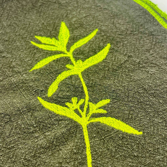 Serviette de table en lin lavé de couleur vert safari, ornée de finitions en jaune néon. Elle présente un motif brodé de sarriette, une plante aromatique. Vue du détail de la broderie.