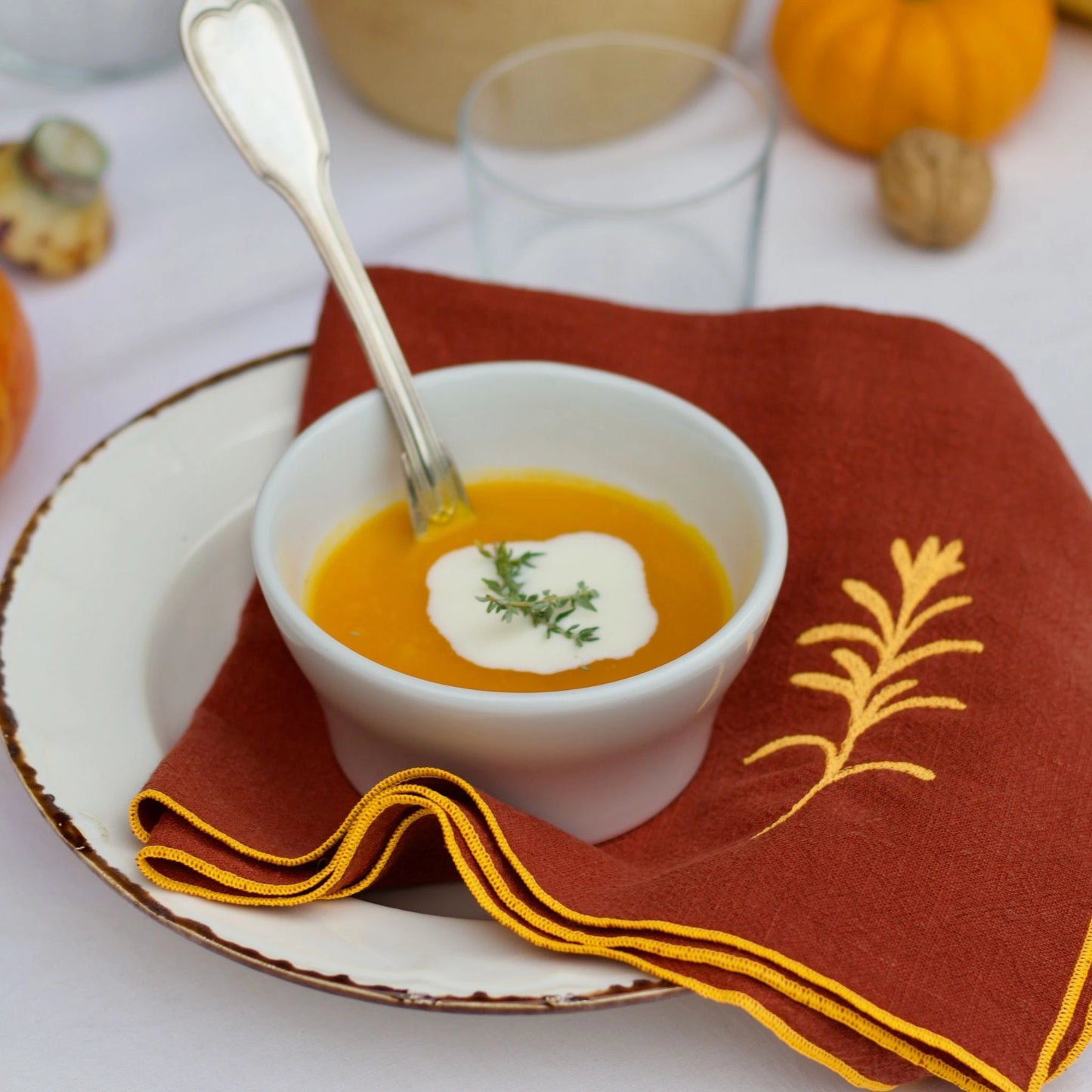 Serviette de table en lin lavé de couleur piment, ornée de finitions en orange néon. Elle présente un motif brodé de romarin, une plante aromatique. Vue du produit en situation.