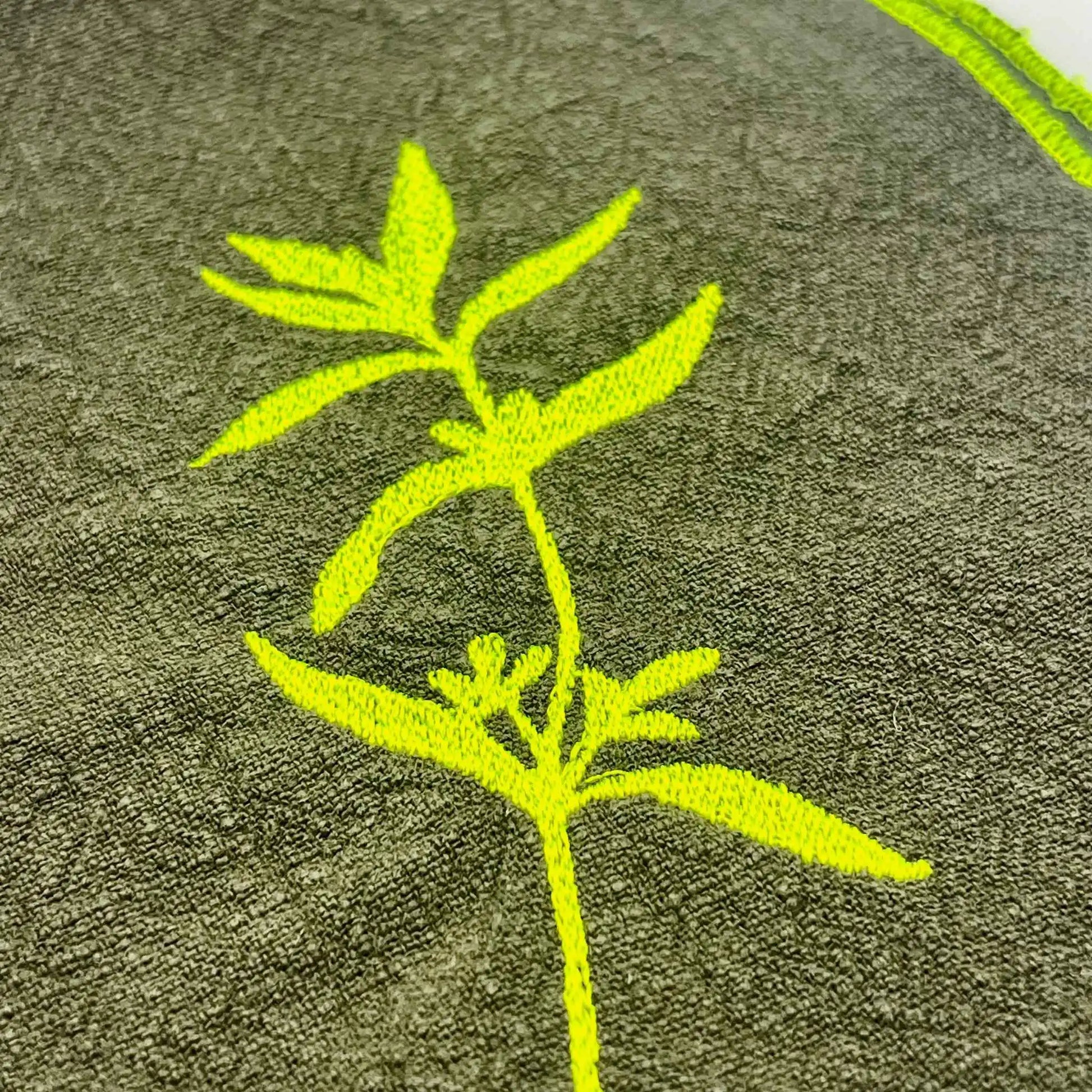 Serviette de table en lin lavé de couleur vert safari, ornée de finitions en jaune néon. Elle présente un motif brodé de sarriette, une plante aromatique. Vue du détail de la broderie.